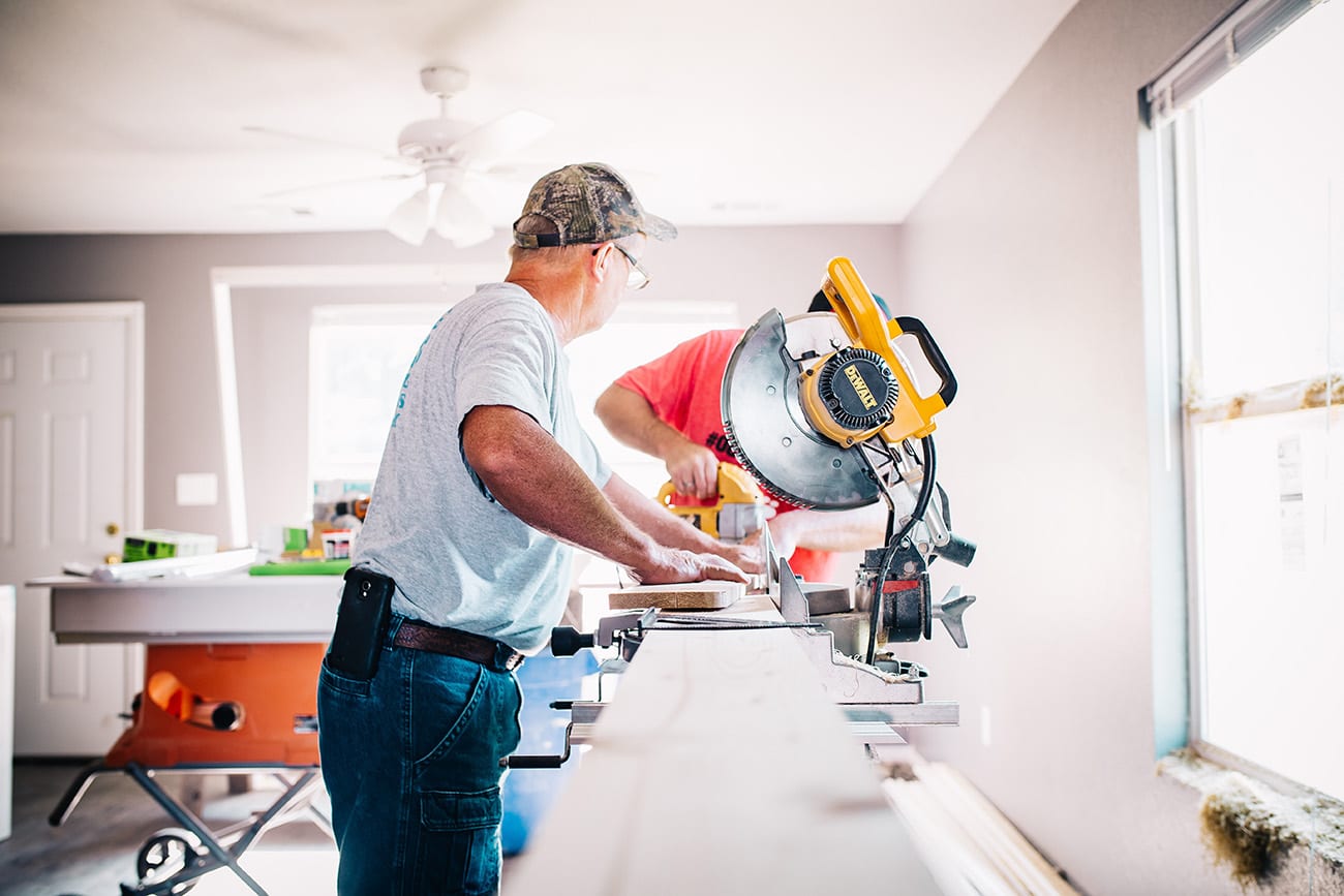 Two people using power tools to cut wood inside a sunlit room under renovation. One person in a gray shirt and cap positions the wood, while the other in a red shirt operates a yellow miter saw.