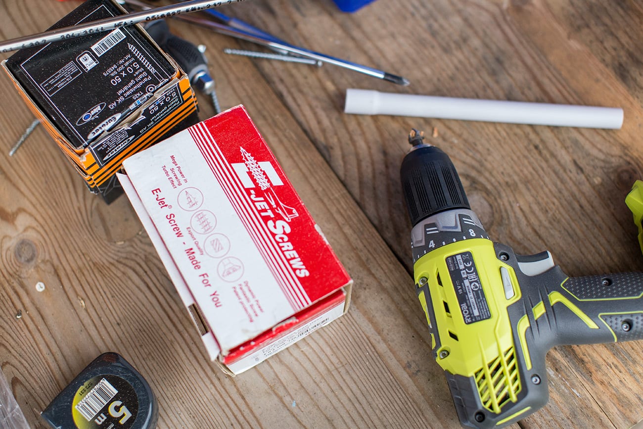 A yellow and black cordless drill, a red and white box labeled Jet Screws, and other tools and hardware are scattered on a wooden work surface.