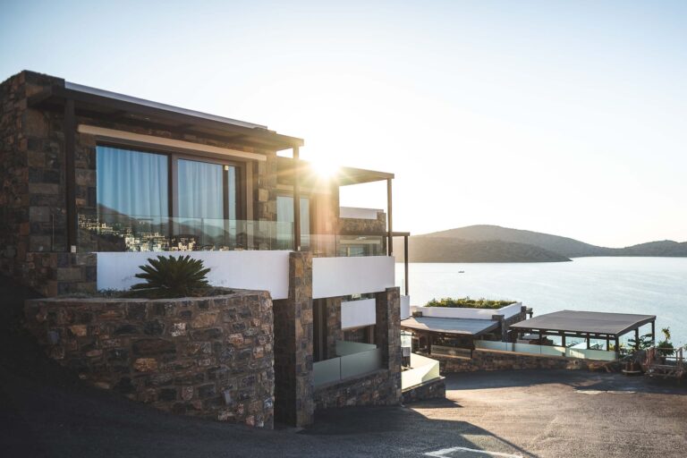 Modern stone and glass house on a hill overlooks a calm, sunlit lake or sea, with mountains in the background and clear skies, creating a tranquil, scenic setting.