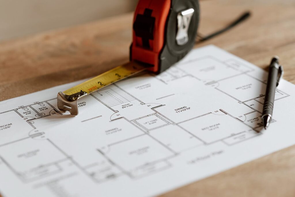 A close-up of a floor plan on a table with a tape measure and a pen resting on top, suggesting planning or measuring for a construction or renovation project.