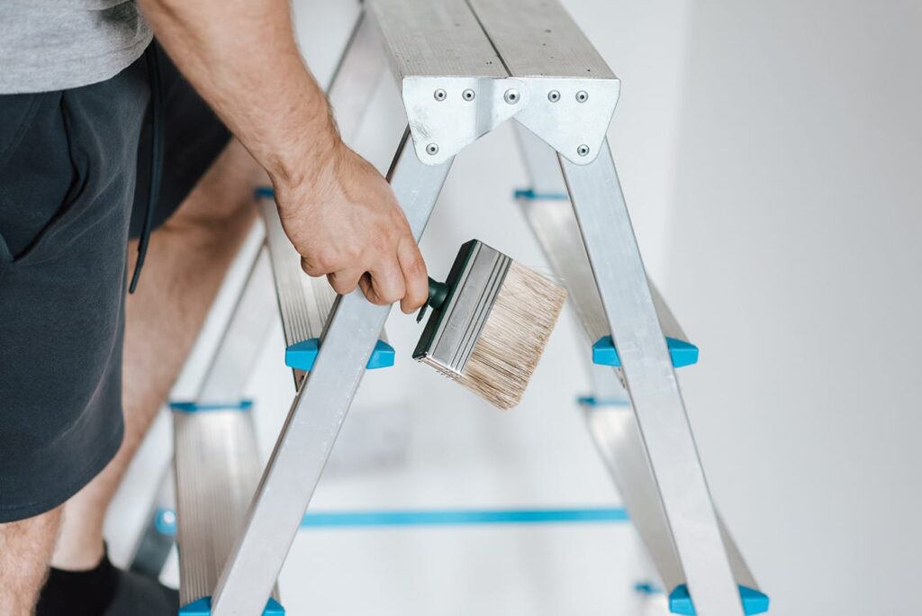 A person in shorts stands on an aluminum stepladder, holding a wide paintbrush with light bristles and a green handle, preparing for painting or home improvement work.