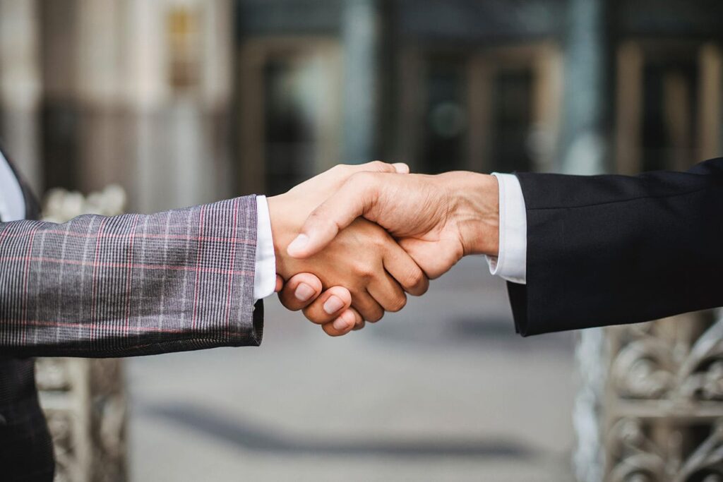 Two people in business attire are shaking hands outdoors, with a blurred background of buildings, symbolizing agreement or partnership.