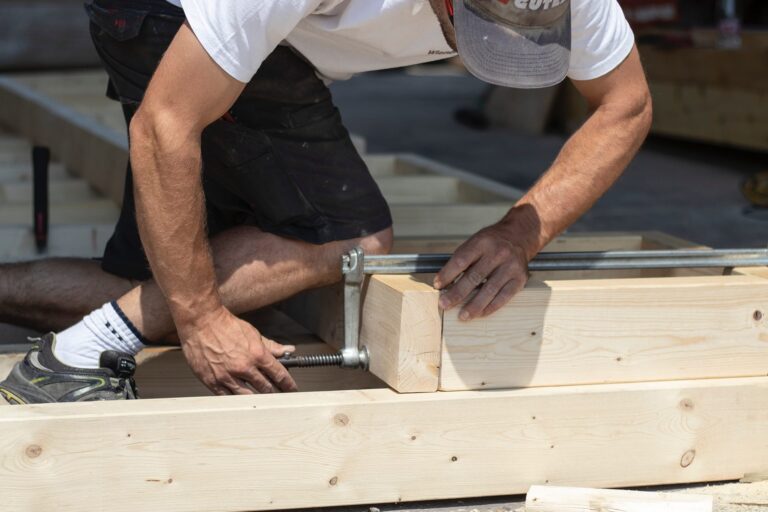 A person in shorts and a cap uses a clamp to secure two pieces of light-colored wood together while kneeling on a wooden frame, working on a construction project.