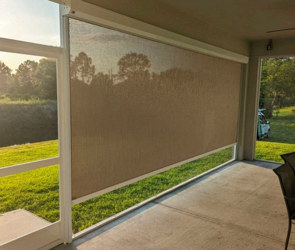 A sunshade is lowered over the open side of a covered patio, filtering sunlight onto the concrete floor. Outside, there is green grass, trees, and a golf cart partially visible in the background.