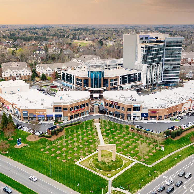 Aerial view of a modern commercial complex with white-roofed buildings, a high-rise tower, parking lots, landscaped green space, and surrounding residential neighborhoods under a cloudy sky.