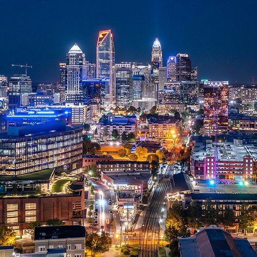 A Charlotte NC skyline at night with tall, illuminated buildings, colorful lights, and busy streets. Railroad tracks run through the foreground, and the scene is filled with urban energy.