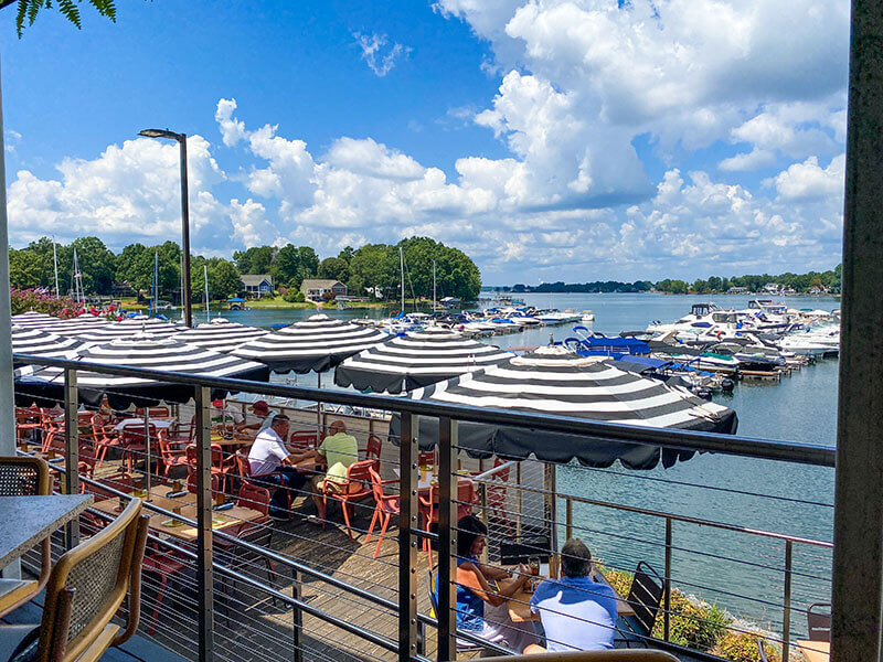 Outdoor dining patio by a lake, with black-and-white striped umbrellas shading tables. People are seated, enjoying food, and several boats are docked nearby under a bright blue sky with scattered clouds.
