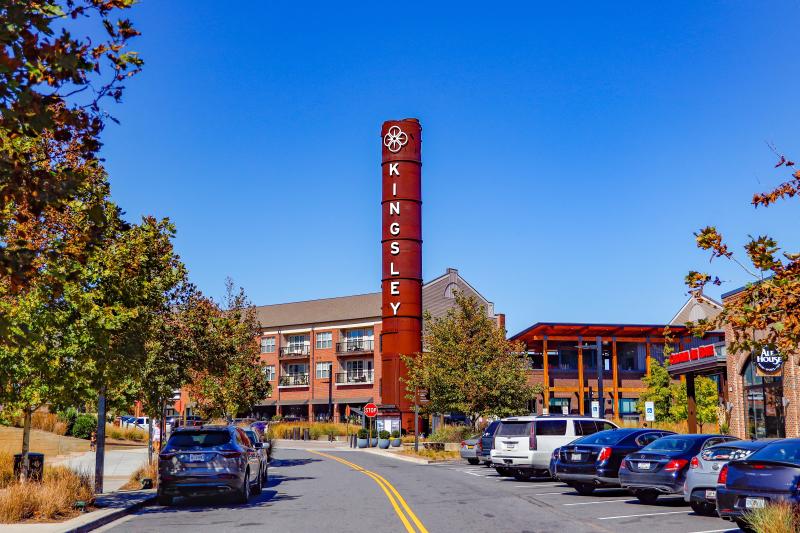 A tall red sign reading KINGSLEY stands in the center of a shopping and residential area with parked cars, trees, and brick buildings under a clear blue sky.