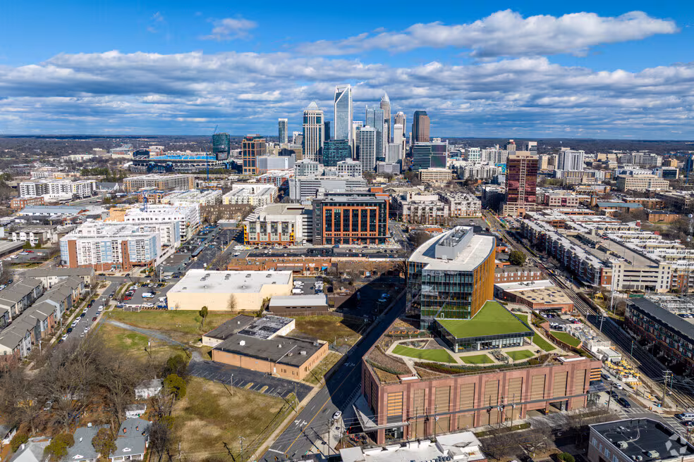 Aerial view of a modern city with tall skyscrapers in the background, mixed-use buildings, streets, and green rooftops in the foreground under a blue sky with scattered clouds.
