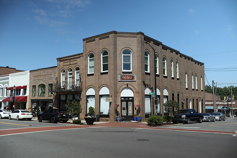 A two-story brick building with arched windows on a sunny day, located on a street corner with parked cars and potted plants near the entrance. Other small businesses line the street.