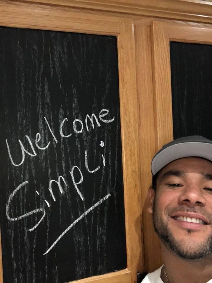Ronald Fletcher wearing a cap smiles in front of a chalkboard with the handwritten message Welcome Simpli on it. The chalkboard is framed with light wood.