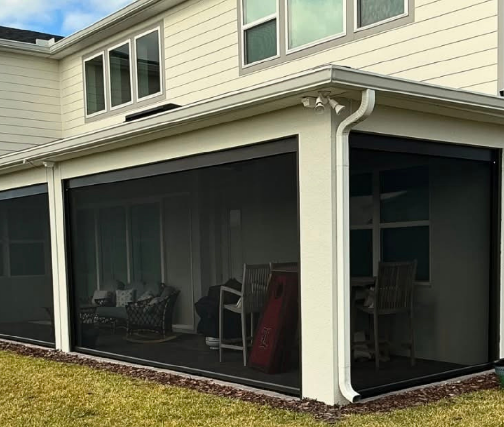 A modern house with a covered patio enclosed by black mesh screens, outdoor furniture inside, and a well-kept lawn in the foreground.