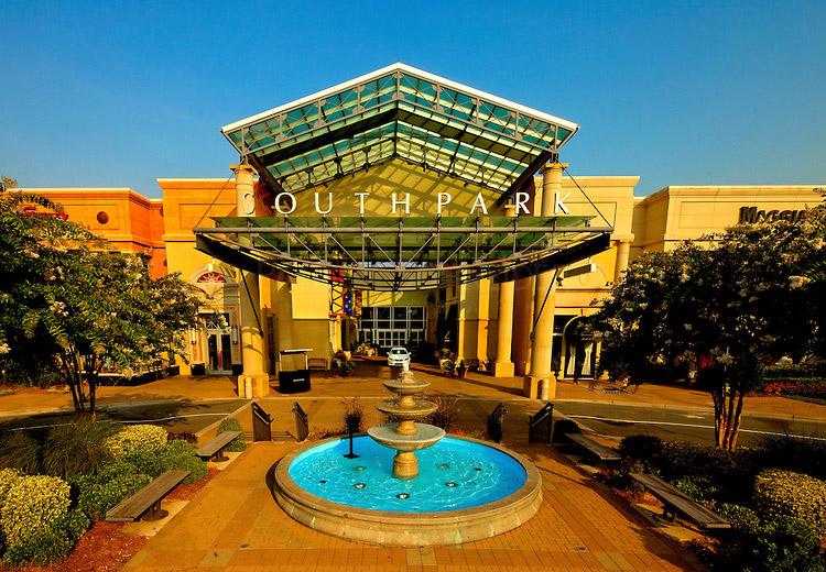 Entrance to SouthPark Mall with a large glass canopy, a three-tiered fountain in front, benches, landscaping, and sunlight casting a warm glow on the building and surroundings.