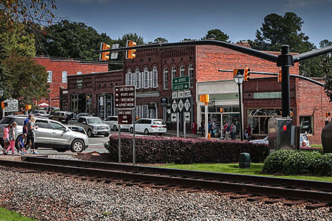 A small town street scene with brick buildings, parked cars, people walking, traffic lights, street signs, and railroad tracks in the foreground surrounded by trees.