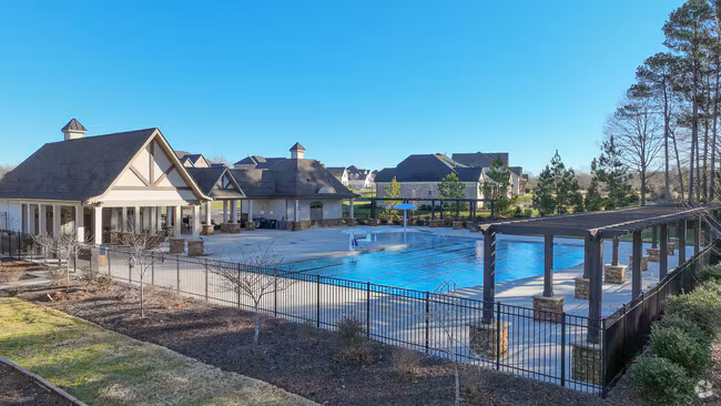 A large outdoor swimming pool with clear blue water is surrounded by a black metal fence, several buildings, and a wooden pergola. Trees and neatly landscaped areas are visible under a clear blue sky.