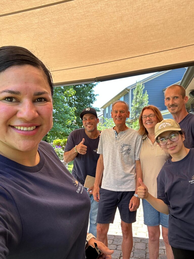 A group of six people, some wearing matching navy shirts and caps with Sunpro Dealer logos, smile and give thumbs up while standing outside on a sunny day near a house and garden.