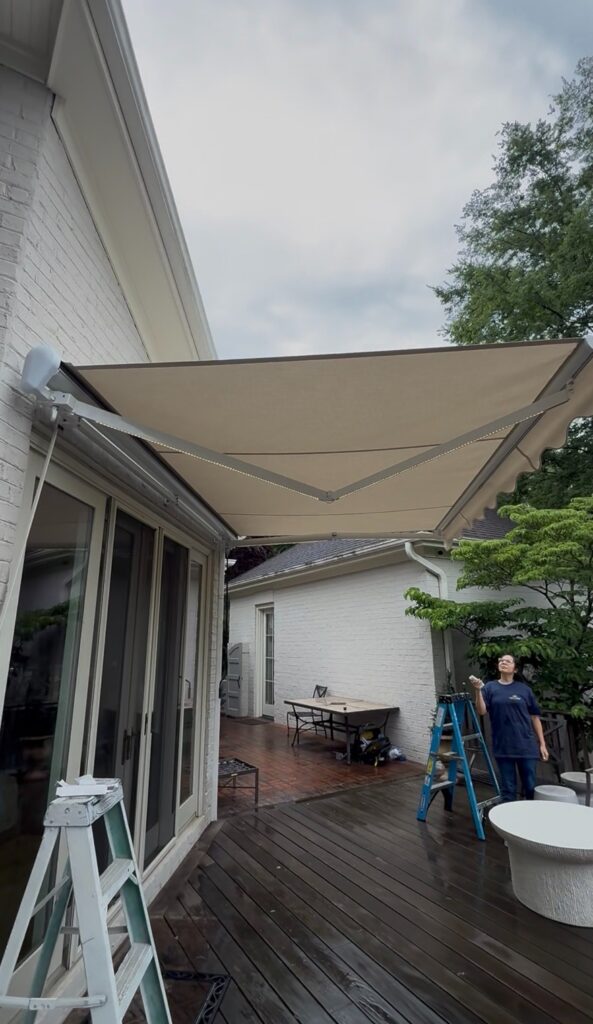 A retractable beige awning, installed by a Sunpro dealer, extends over a wooden deck attached to a white brick house. A person stands on the deck, holding a remote next to a blue ladder. Patio furniture and trees are visible in the background.