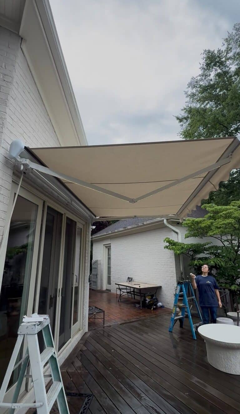 A retractable beige awning, installed by a Sunpro dealer, extends over a wooden deck attached to a white brick house. A person stands on the deck, holding a remote next to a blue ladder. Patio furniture and trees are visible in the background.