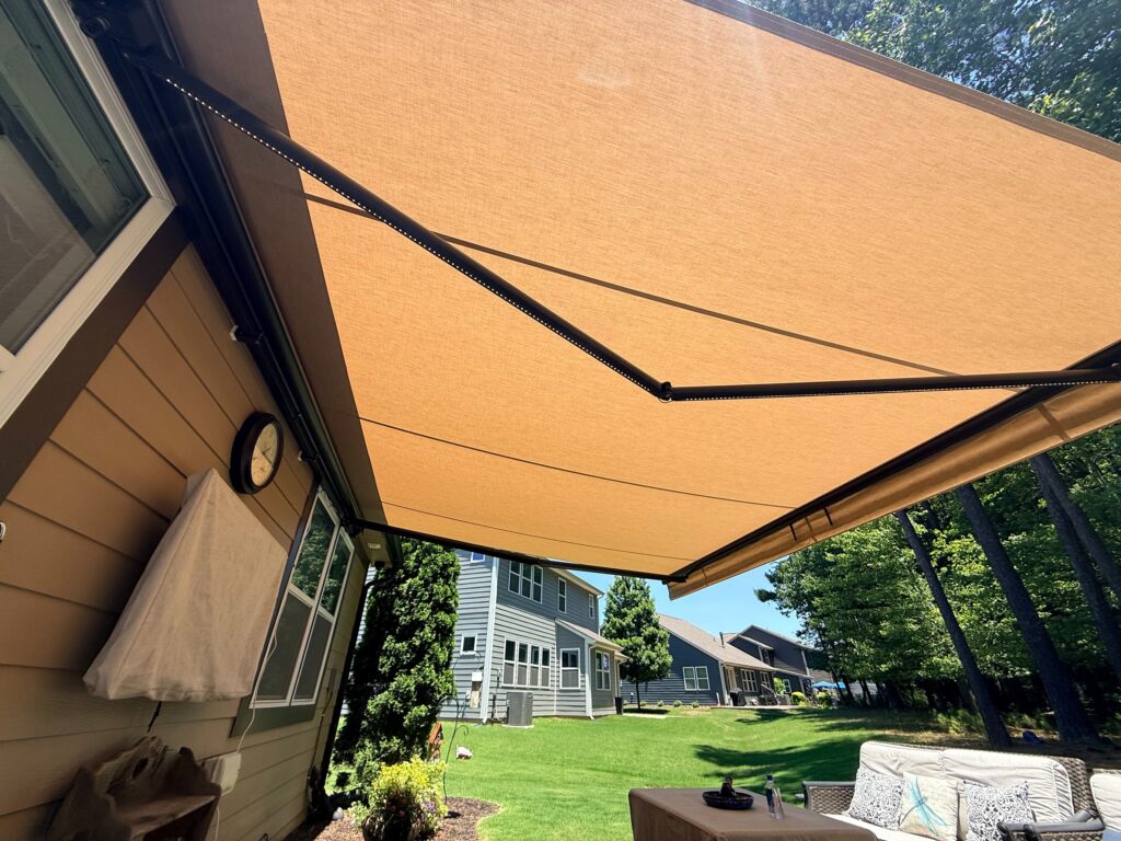 A large tan retractable awning, installed by a SunPro dealer, extends over a backyard patio and shades the seating area. Nearby, a beige house and other homes are visible among green grass and tall trees under a sunny sky.
