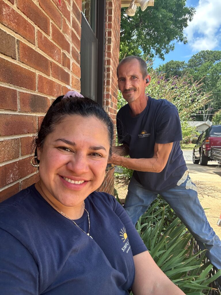 Two people in navy shirts smile outside near a brick wall and green plants on a sunny day, representing their roles as Sunpro dealer team members. A woman is in the foreground, and a man stands behind her. Trees and a red truck are visible in the background.
