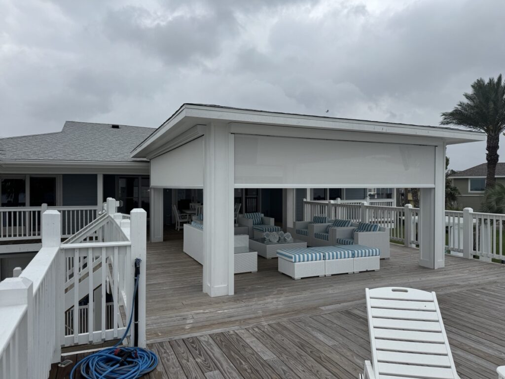 A covered outdoor seating area with white and blue cushioned chairs sits on a wooden deck. An overcast sky looms above, and a palm tree is visible in the background. A hose lies coiled nearby.