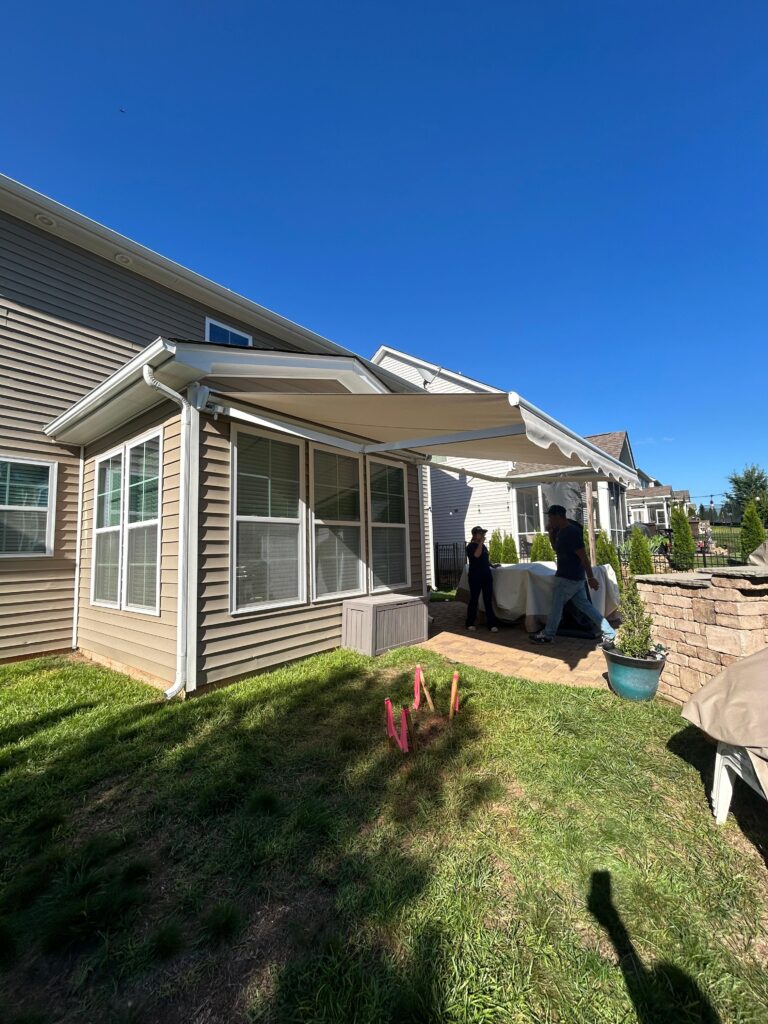 Two people stand on a brick patio under a large, extended Sunpro dealer awning attached to a beige house on a sunny day. The lawn is green, with pink survey flags and a potted plant near a stone retaining wall.