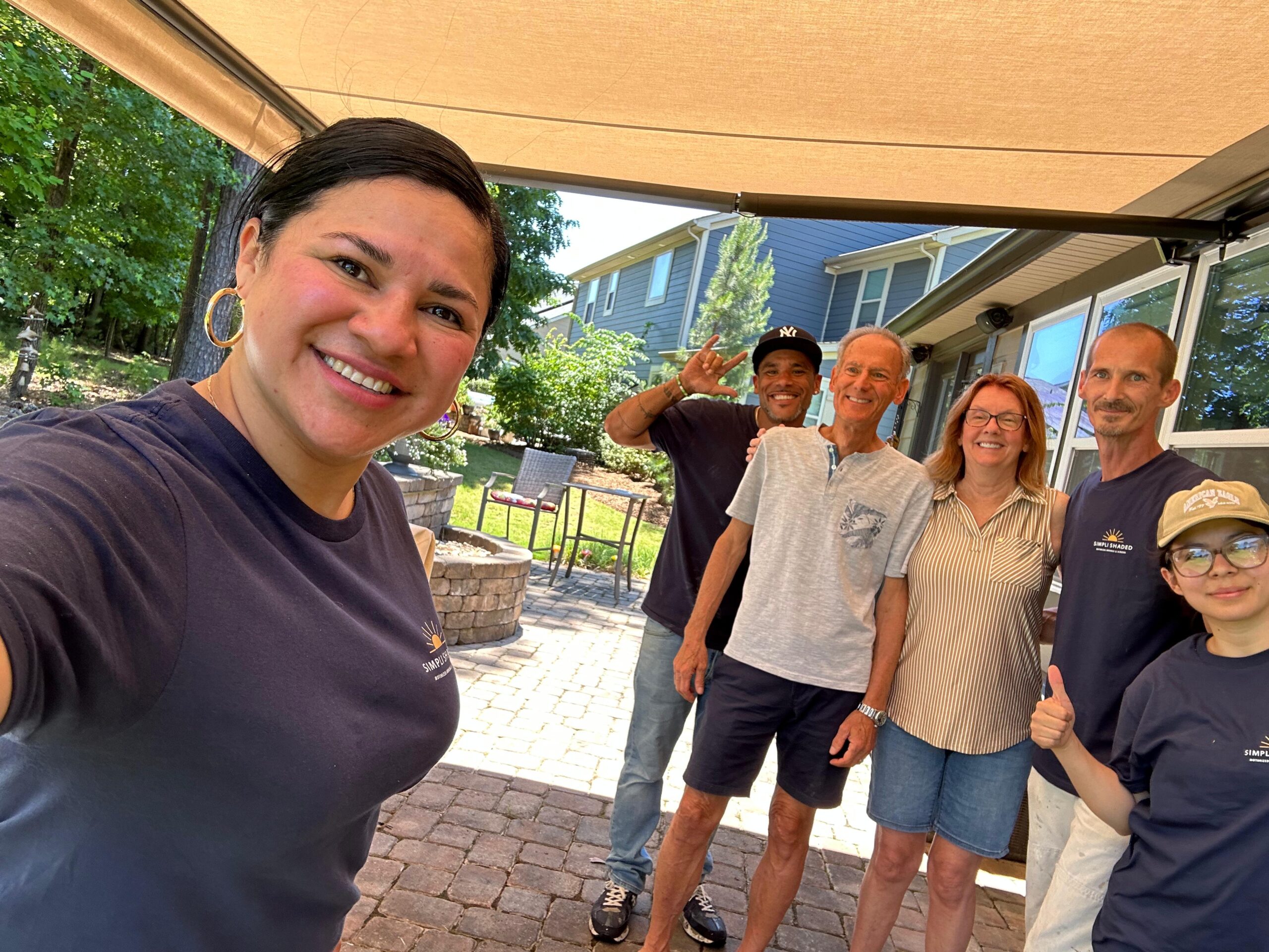 Six people, possibly part of a Sunpro dealer team, smile for a group selfie outdoors under a canopy, with trees and a house in the background. Most are wearing navy shirts, and one person gives a thumbs up while another flashes a peace sign.
