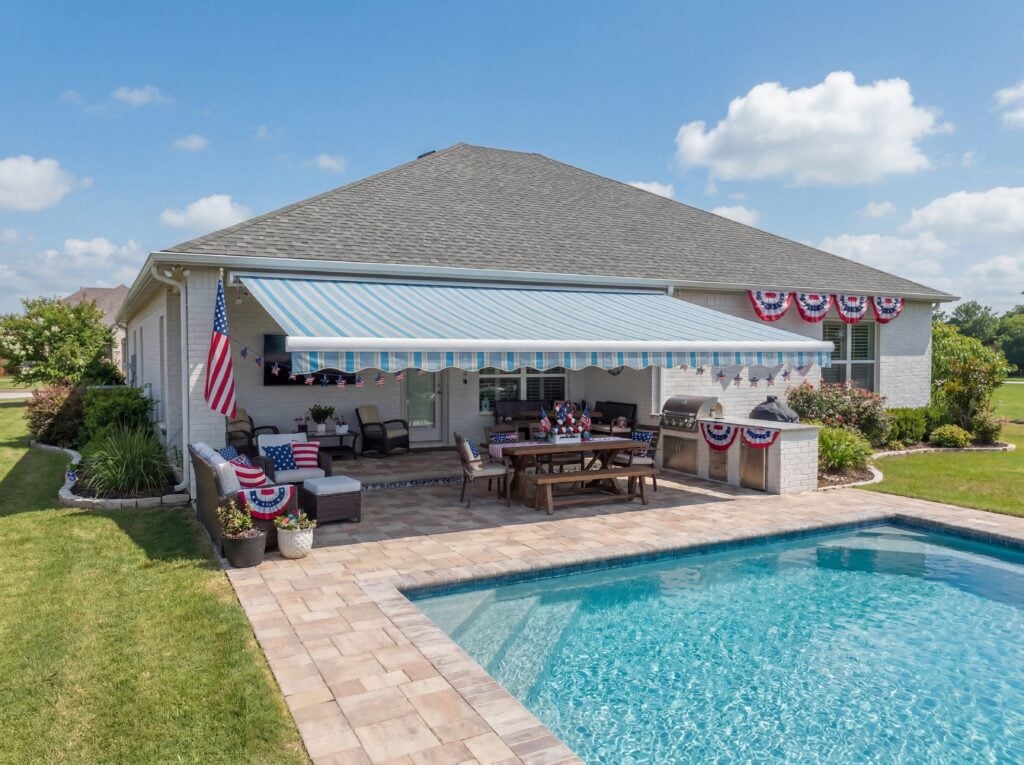 A backyard patio with a pool, outdoor dining set, grill, and a blue-and-white striped awning showcases the expertise of a trusted SunPro dealer. Patriotic red, white, and blue decorations adorn the house and patio, while the sky is partly cloudy.