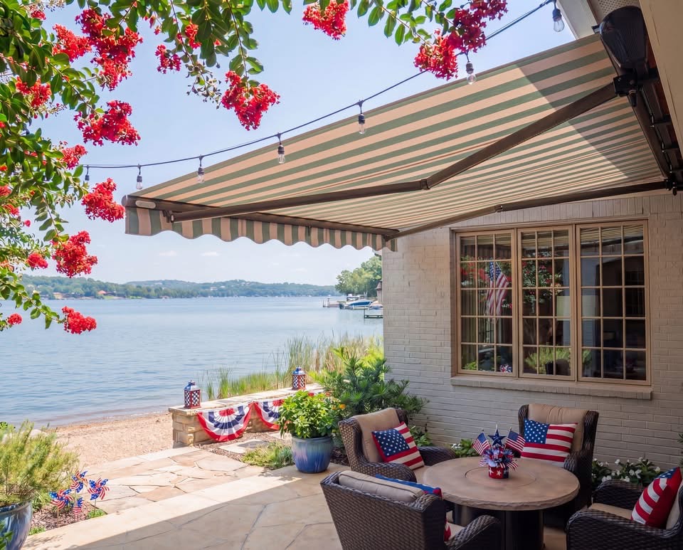 A lakeside patio with a striped awning, outdoor chairs with American flag pillows, festive red flowers, and patriotic decor—expertly installed by your local SunPro dealer. The view overlooks calm water and distant trees under a sunny sky.