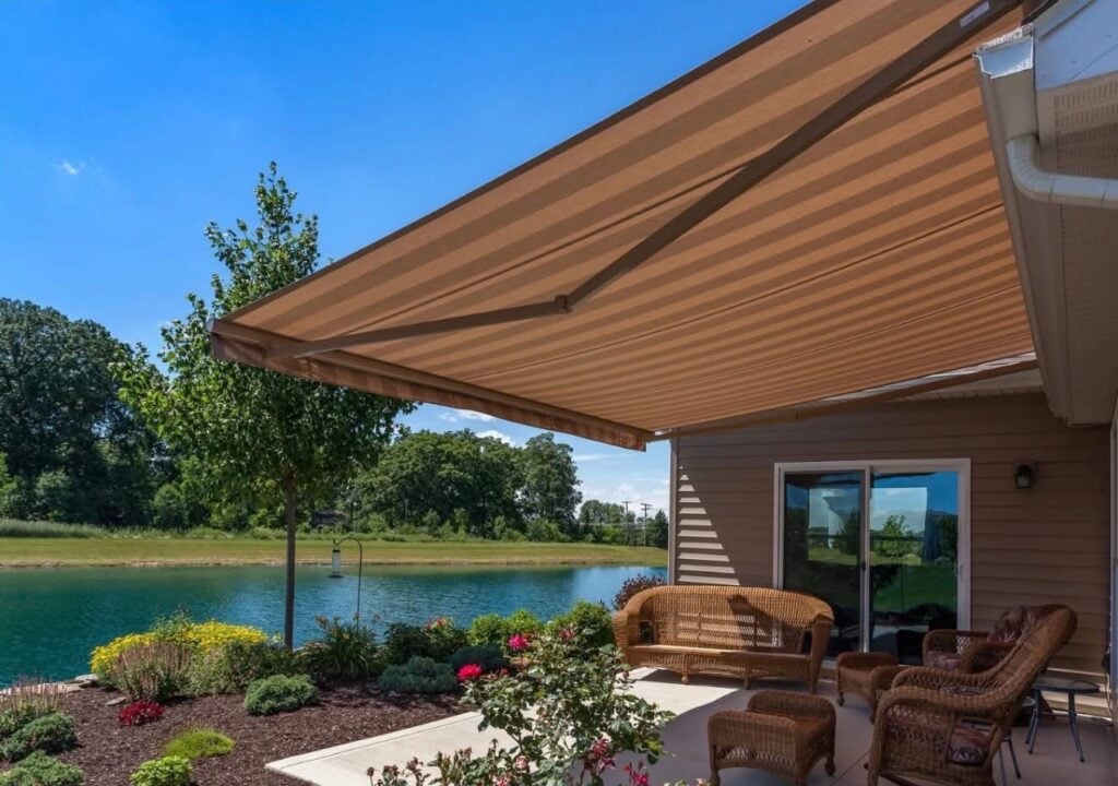 A striped retractable awning, expertly installed by a local sunpro dealer, extends over a patio with wicker furniture, potted plants, and flowers, beside a small pond and landscaped garden on a sunny day.
