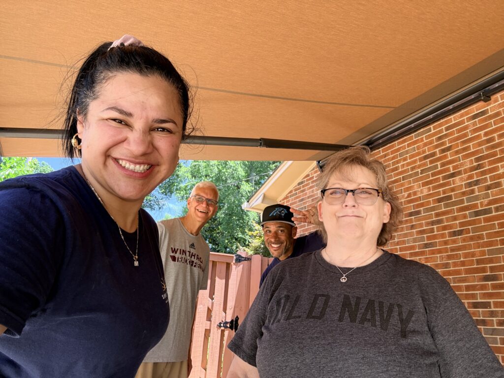 Four people pose and smile outside under a canopy. The group stands near a brick wall and a wooden fence, enjoying a sunny day together with their trusted Sunpro dealer.