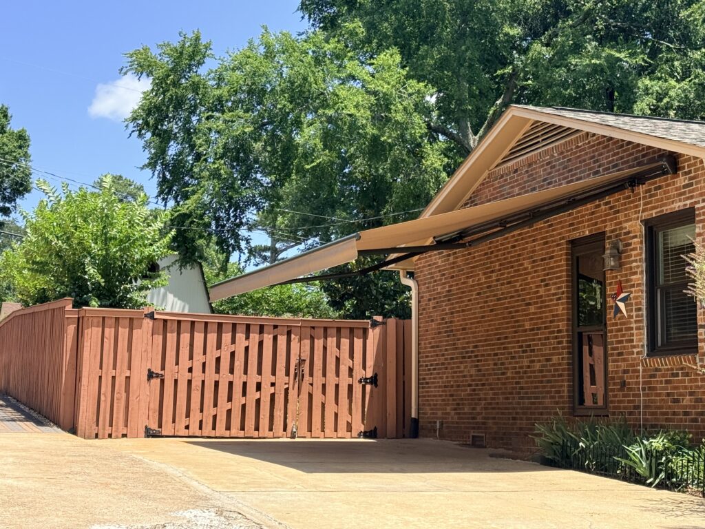A tan retractable awning, installed by a Sunpro dealer, extends over a driveway next to a red brick house with a wooden fence and gate, under a clear blue sky and green trees.
