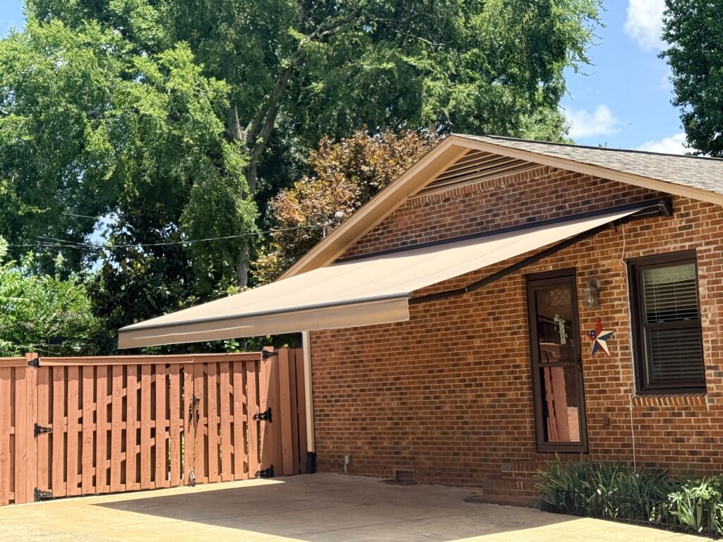 A brick house with a brown retractable awning, expertly installed by a SunPro dealer, extends over a door next to a wooden gate and is surrounded by green trees on a sunny day.