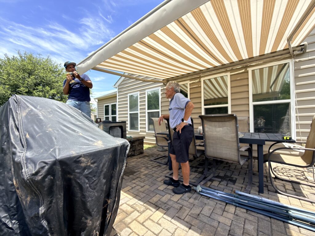 Two people talk on a patio under a striped awning from a local SunPro dealer. One stands on the ground, the other on a raised surface. Patio furniture and a covered grill are visible. It’s a bright, sunny day with blue sky and some clouds.