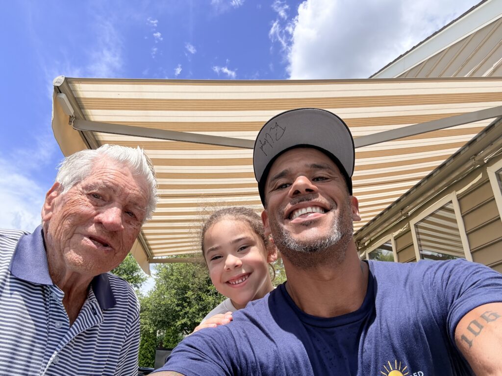 Three people—an older man, a young girl, and a middle-aged man—smile outdoors under a striped awning provided by their trusted Sunpro dealer. The sky is blue with scattered clouds, and the middle-aged man wears a hat and a blue shirt.