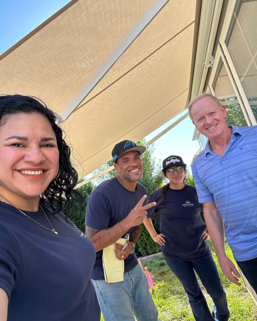Four adults smiling under a sunshade outdoors; one woman holds the camera for a selfie, while the man next to her—likely a SunPro dealer—makes a peace sign. Two others stand near a building with glass windows on this sunny day.