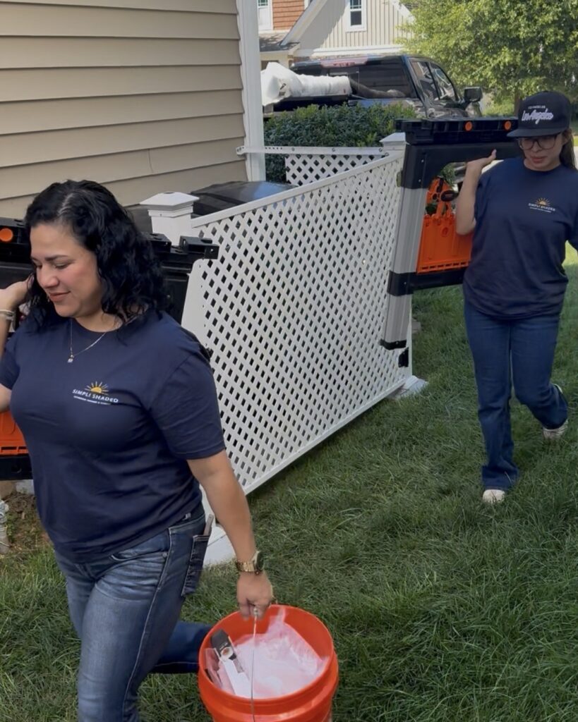 Two women in navy shirts and jeans, both sunpro dealers, carry tools and a bucket outside near a house and white lattice fence on a grassy lawn.