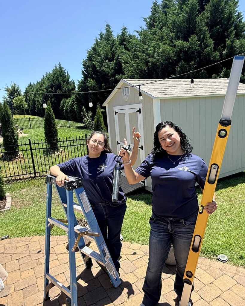 Two women in blue shirts stand outside near a shed, showcasing their skills as sunpro dealers. One holds a level while the other stands confidently on a ladder. Both are smiling, with tools in hand, and string lights hang above them. It’s a sunny day, with green grass and trees in the background.