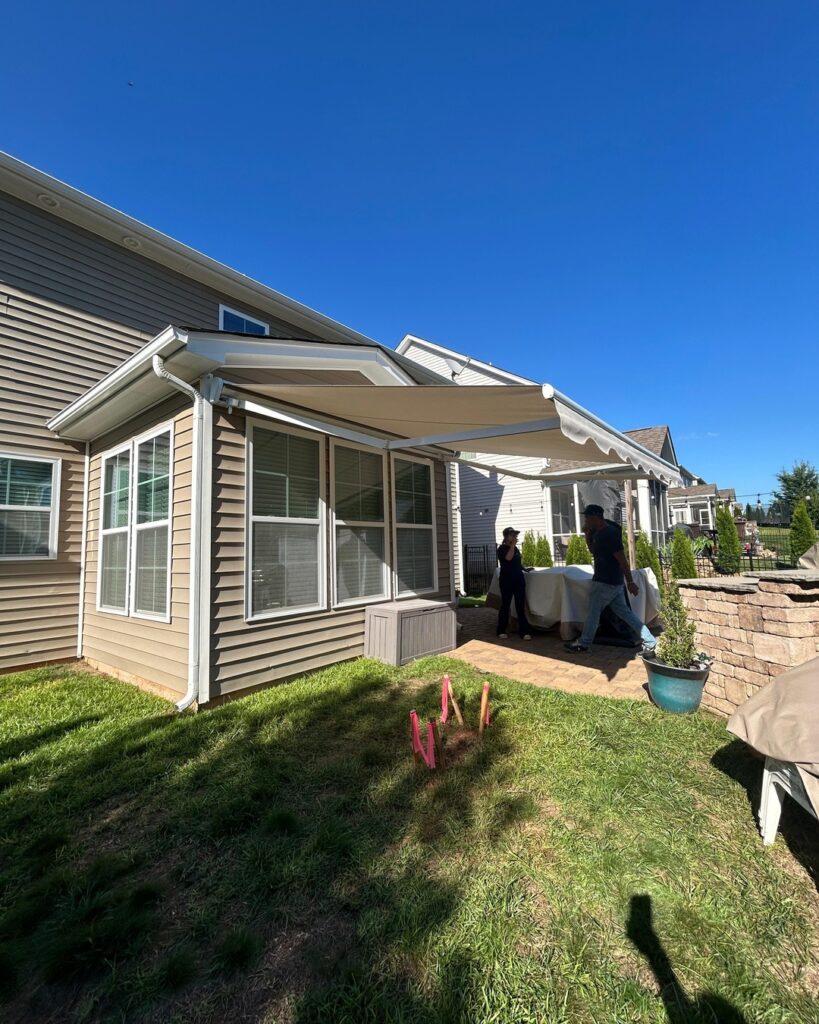 A retractable awning extends over a patio outside a tan house, showcasing the quality you’d expect from a trusted SunPro dealer. Two people stand near a table under the awning, enjoying the shade on a clear, blue day. Pink lawn markers are visible on the grass.