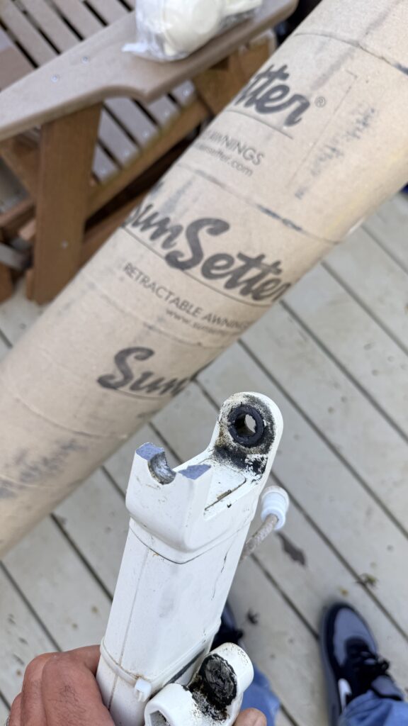 A hand holds a broken white metal bracket near a rolled-up SunSetter retractable awning on a wooden deck. As a SunPro dealer might observe, the setup includes a wooden chair and shoes visible in the background.