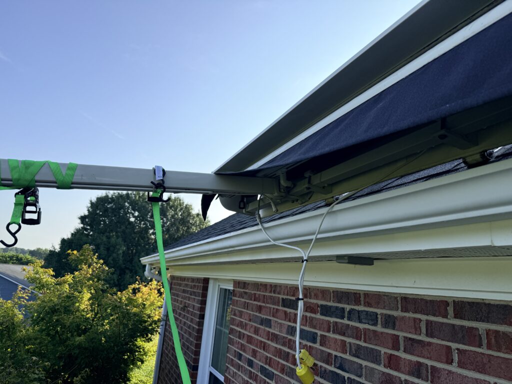 Close-up of a brick house with a green strap and metal pole attached to the roof gutter, electrical wires hanging down, and trees in the background under a clear blue sky—an ideal setting for a Sunpro dealer to showcase solar solutions.