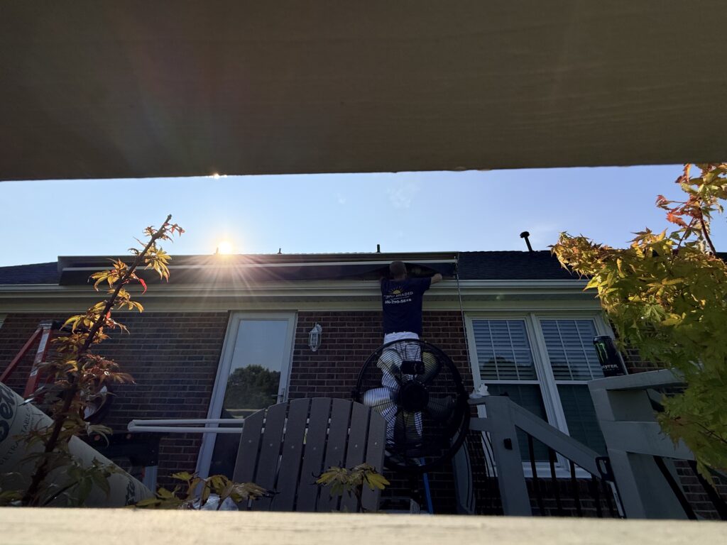 A person stands on a ladder fixing a roof gutter on a brick house in bright sunlight. Patio furniture, plants, and a large fan are visible in the foreground, creating an inviting outdoor space that highlights the quality workmanship you’d expect from a Sunpro dealer.
