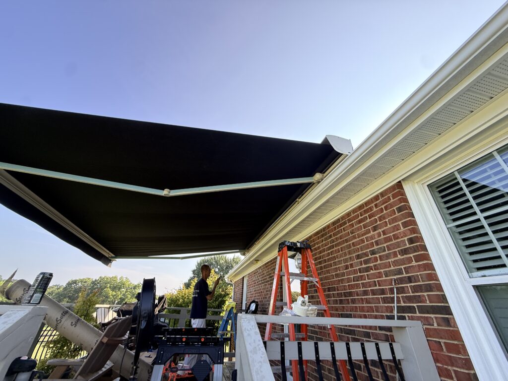 A black retractable awning, installed by a Sunpro dealer, is extended over a deck. A person stands near a red ladder, and tools are scattered on the deck. The house has brick siding and a white window with blinds. The sky is clear and blue.