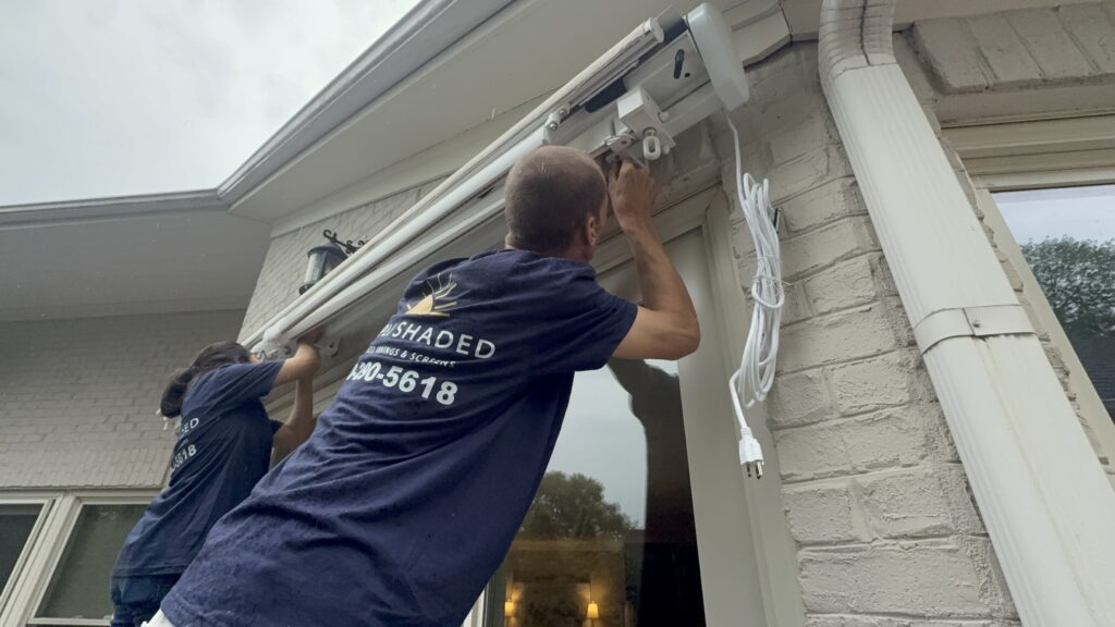 Two people in navy shirts, representing a SunPro dealer, install an awning on the exterior wall of a house. One person is adjusting the awning’s bracket near a glass door, while the other assists. A phone number is visible on the back of one shirt.