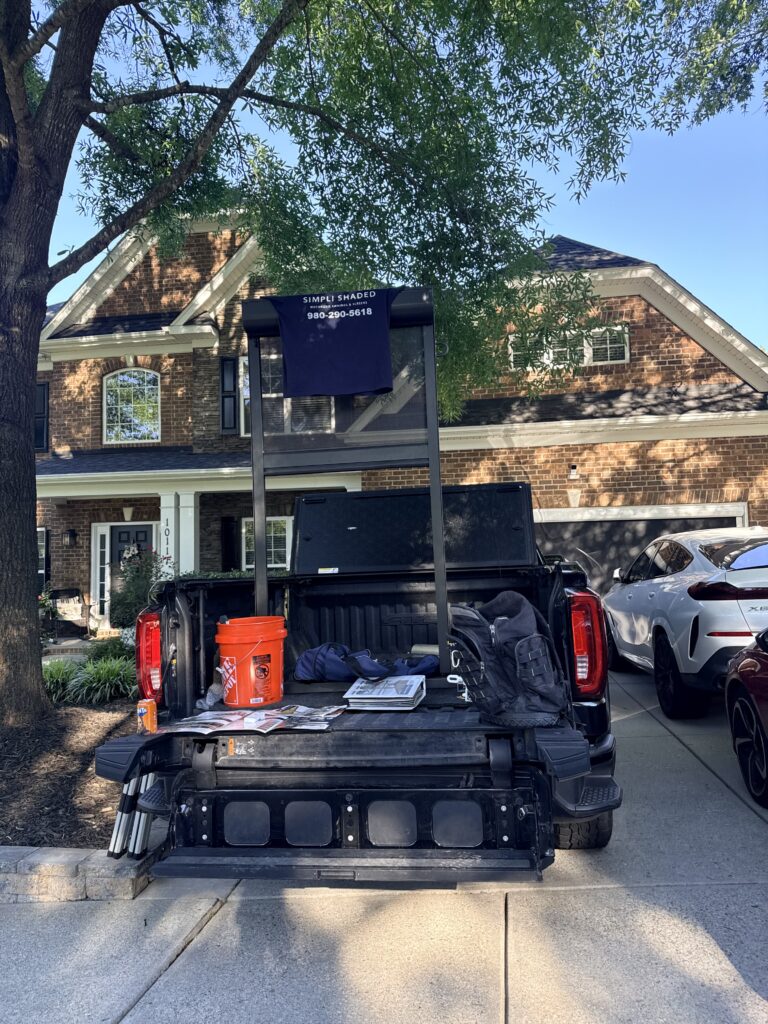 A pickup truck with its tailgate down displays tools, a shirt on a stand, and buckets in the bed—giving the impression that a Sunpro dealer is ready for work. The truck is parked in a driveway in front of a brick house, with trees providing shade.