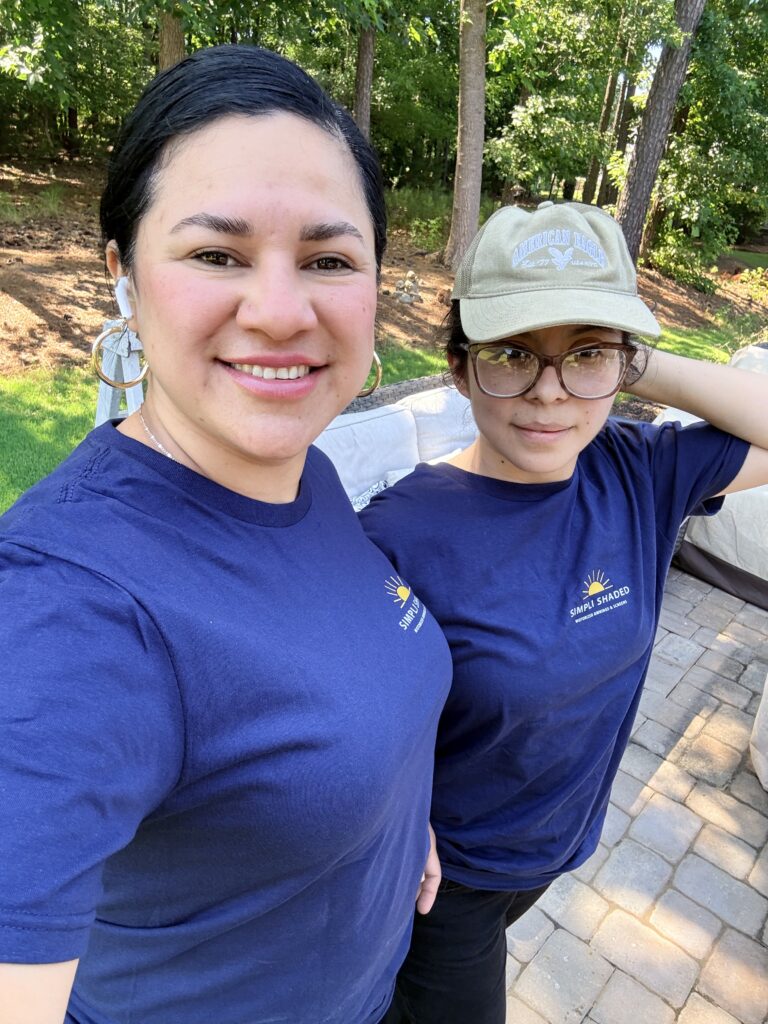 Two people stand outdoors on a sunny day, smiling at the camera. Both wear navy blue shirts—one sporting a beige cap and glasses, giving off a friendly sunpro dealer vibe. Trees and a patio are visible in the background.