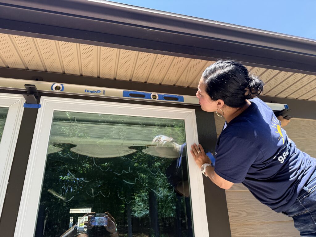 A woman in a navy shirt, likely a SunPro dealer, checks the alignment of a window with a level tool on a sunny day. Trees are reflected in the glass.