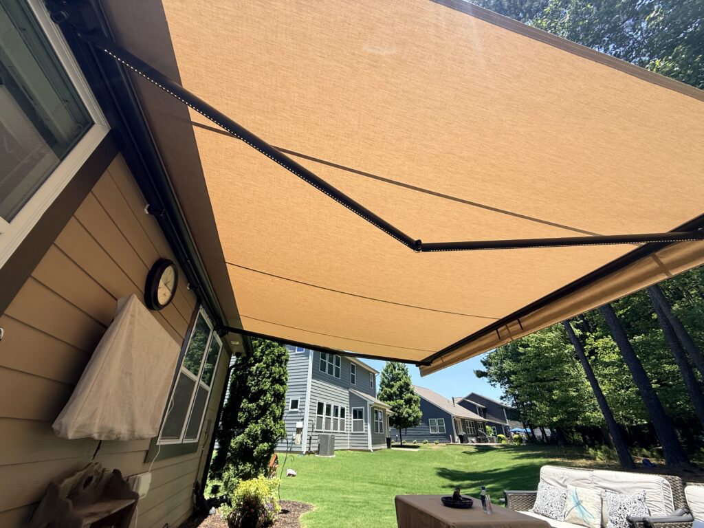 A tan retractable awning extends over a patio beside a house, providing shade, expertly installed by a trusted SunPro dealer. Nearby are houses, green grass, and trees under a clear blue sky.
