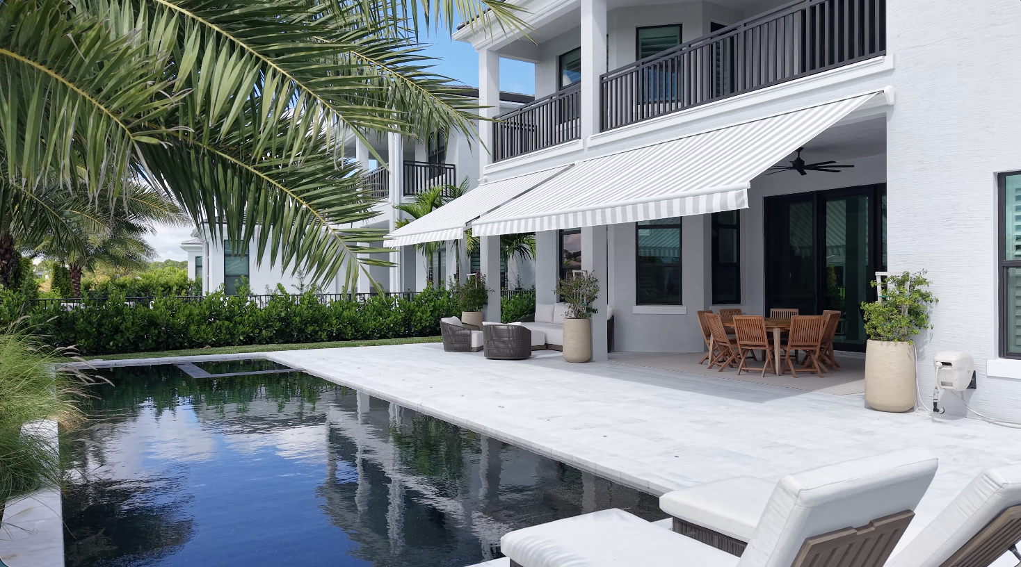 Modern white house with large windows, striped awning, patio furniture, and a swimming pool surrounded by palm trees and greenery under a blue sky.
