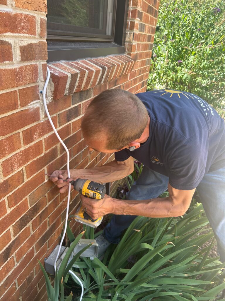 A Sunpro dealer in a blue shirt uses a yellow drill to install a white cable into a brick wall outside near some green plants.